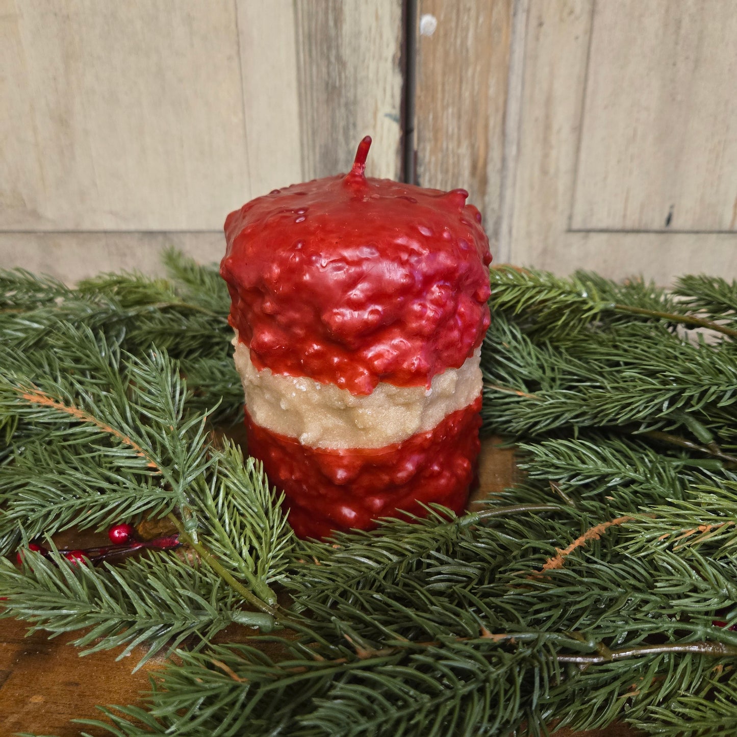 A short pillar candle with a ribbon candy effect, featuring alternating layers of red and white wax, placed on a wooden surface with pine branch decorations around it.