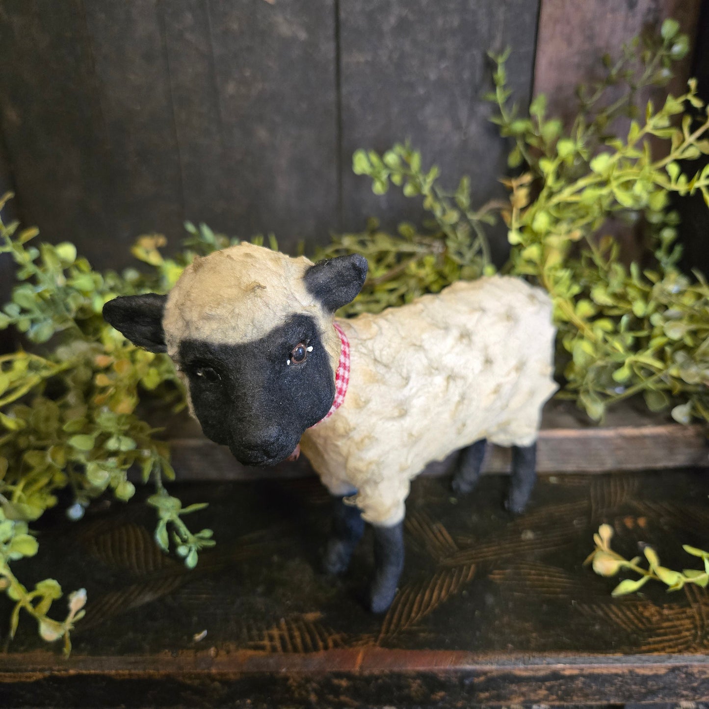 Plush sheep toy with black face and white wool, hanging from a wooden frame with greenery.