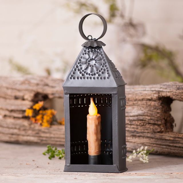 A smokey black finish metal candle lantern with a lit candle inside, placed on a wooden surface with natural decor around it.