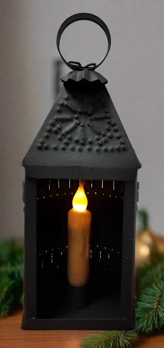 A black textured metal candle lantern with a candle lit inside, designed for use with a 4-inch taper candle (not included).