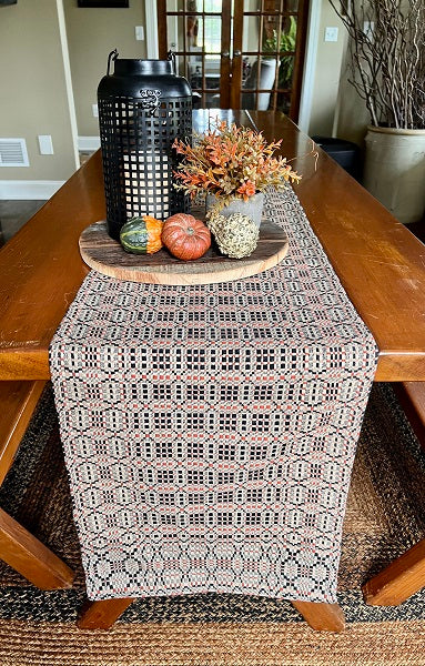 A place mat with a geometric pattern featuring black, rust, and linen colors, displayed on a wooden dining table with a decorative object on top and a vase with flowers in the background.