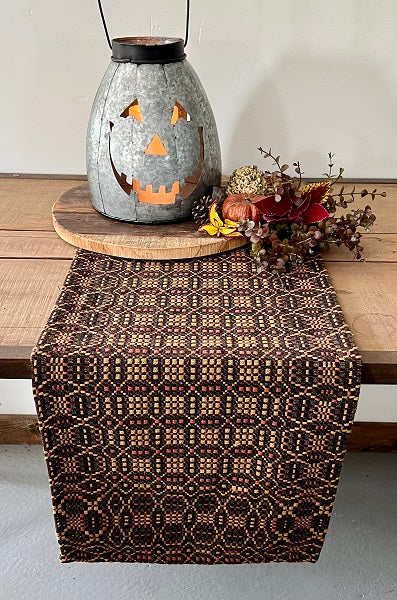 A rectangular placemat with a mustard yellow, black, and pumpkin design, displayed on a wooden dining table with a decorative pumpkin and autumn leaves.