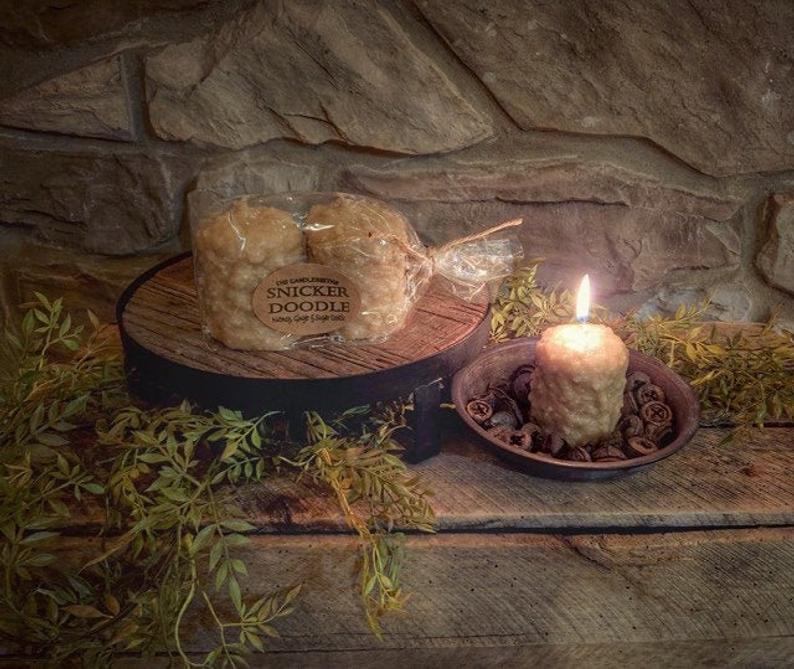 A pack of two Snickerdoodle-scented votive candles, shaped like cookies, displayed on a wooden surface with a faux snickerdoodle finish.