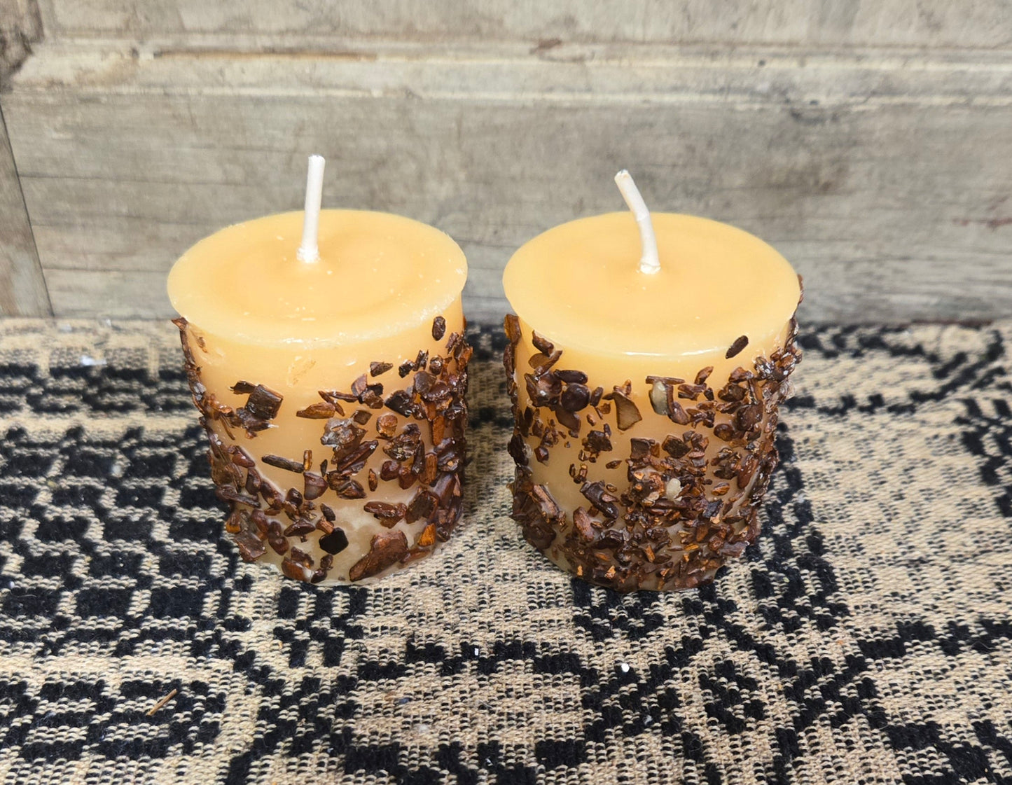 Two snickerdoodle scented votive candles with a warm glow, placed on a textured black mat, with a wooden background.