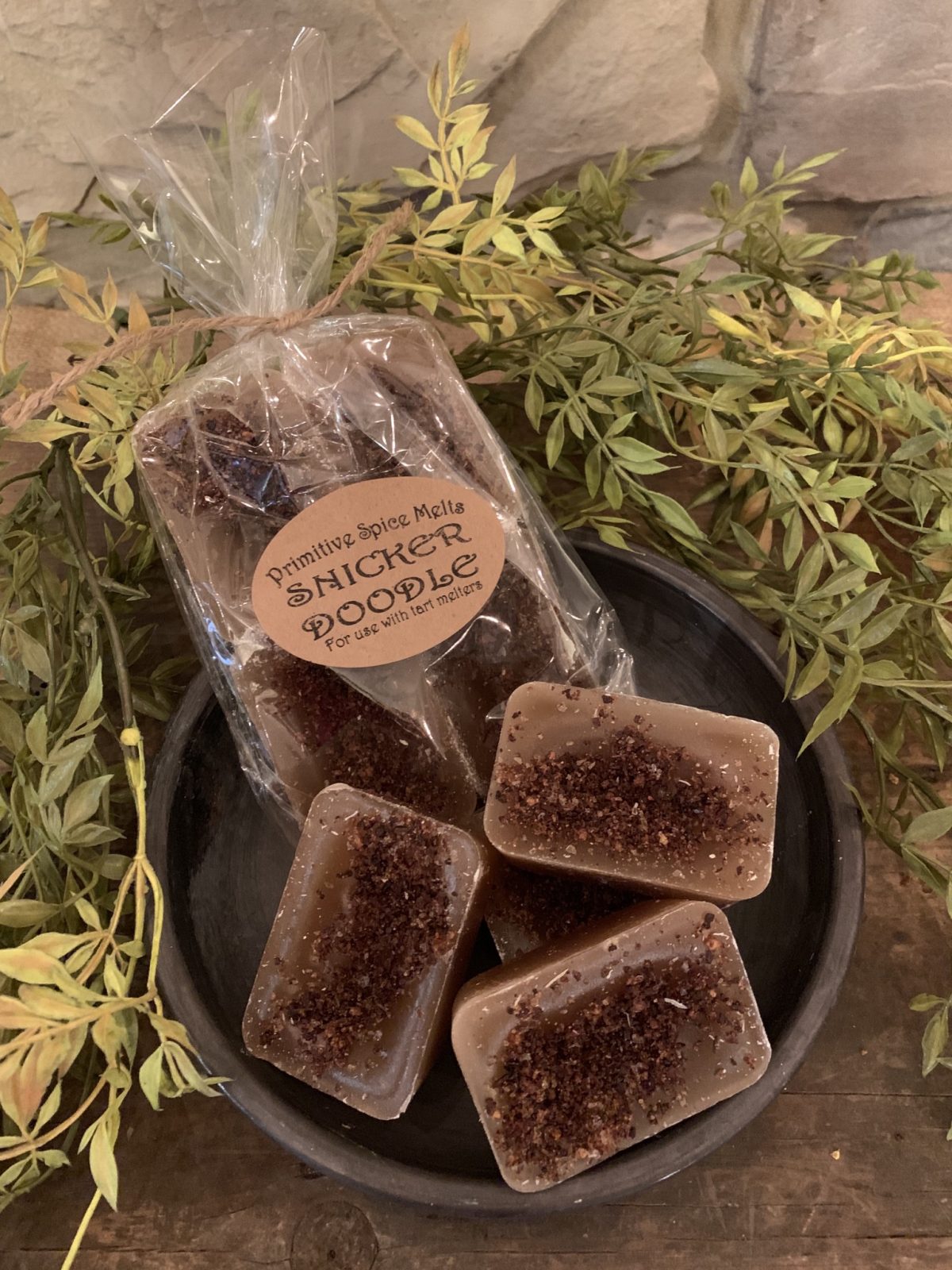 A pack of brown Snickerdoodle wax melts with a label, placed on a black dish with green leaves in the background.