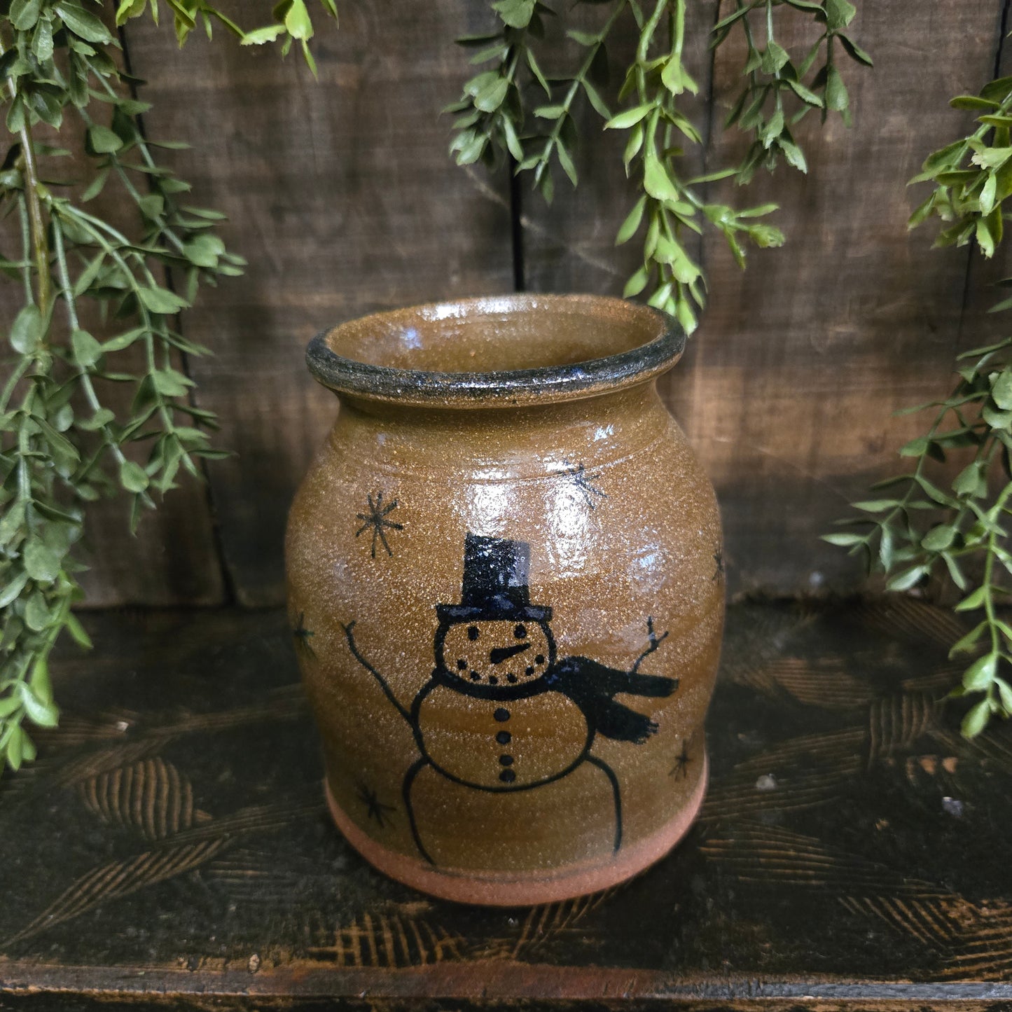 Stoneware crock with a snowman design on a wooden surface with green leaves.