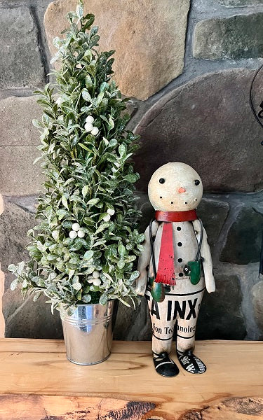 A decorative snowman figure with mittens, placed next to a artificial greenery in a silver pot, on a wooden surface with a stone wall background.