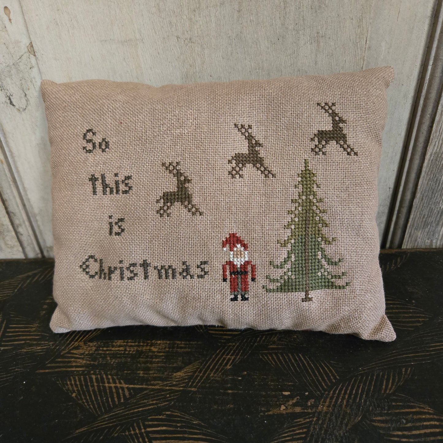 Decorative pillow with cross-stitched design featuring reindeer and 'So this is Christmas' text, placed against a textured wall.