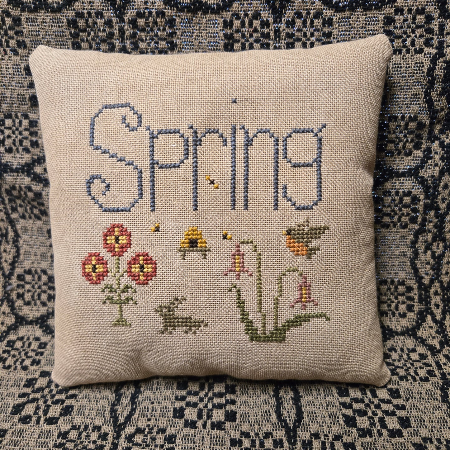 Decorative pillow with cross-stitched design of 'Spring' and floral elements on a patterned background.