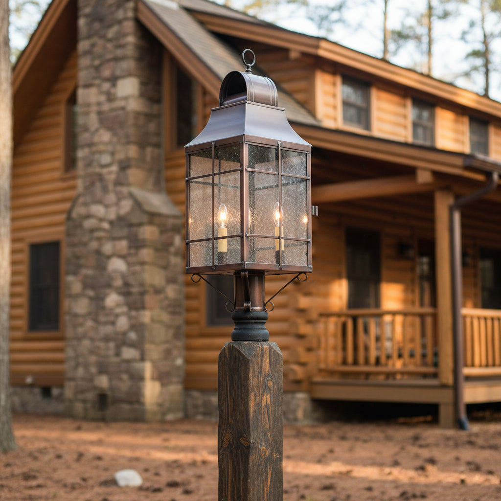 Decorative outdoor lantern with three lit candles mounted on a post in front of log cabin home.
