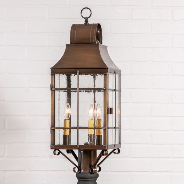 Colonial three light outdoor post light in weathered brass with bubbly glass on white background.