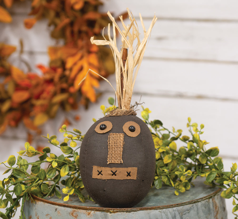 A stuffed toy resembling a black pumpkin with a stitched patchwork face and moss detailing, placed on a blue surface with autumn leaves around it.