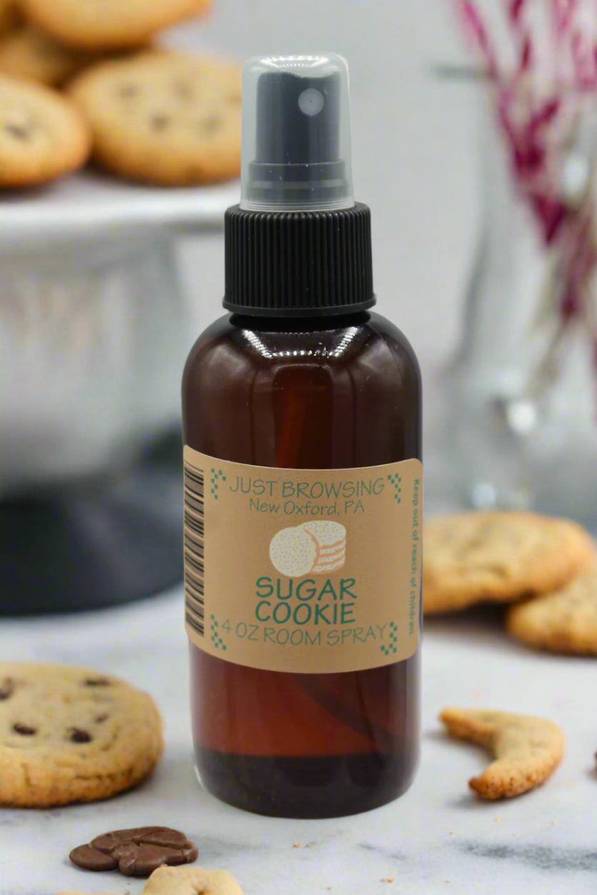 A 4oz bottle of Sugar Cookie room spray with a mist spray top. The bottle is brown with a label that has the product name and an image of a cookie.