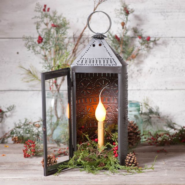 Decorative lantern with a lit candle inside, surrounded by greenery and pinecones on a wooden surface.