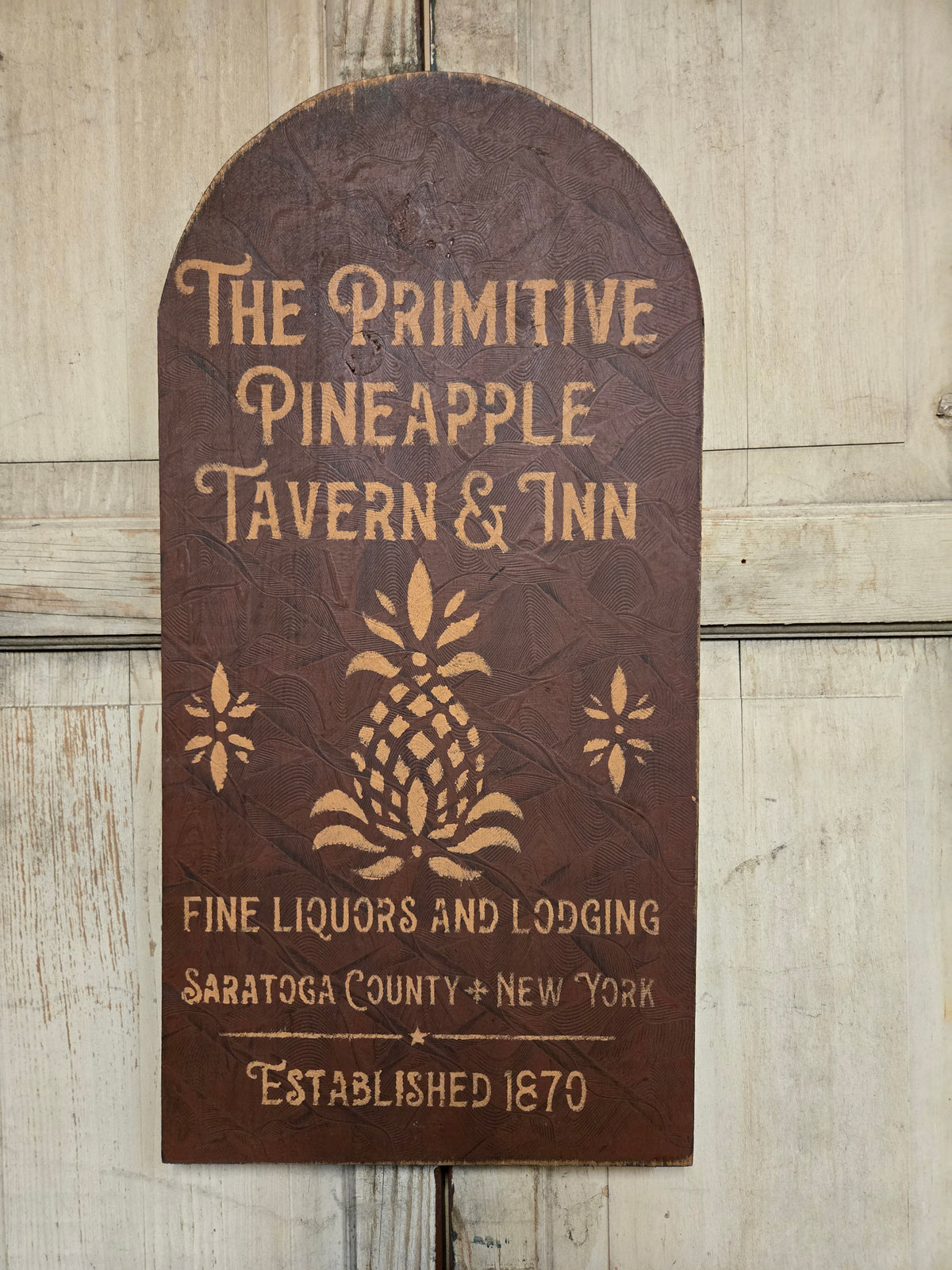 A rustic arched wooden sign for 'The Primitive Pineapple Tavern & Inn' with raised tan lettering and a pineapple motif, mounted against a light wood plank wall.