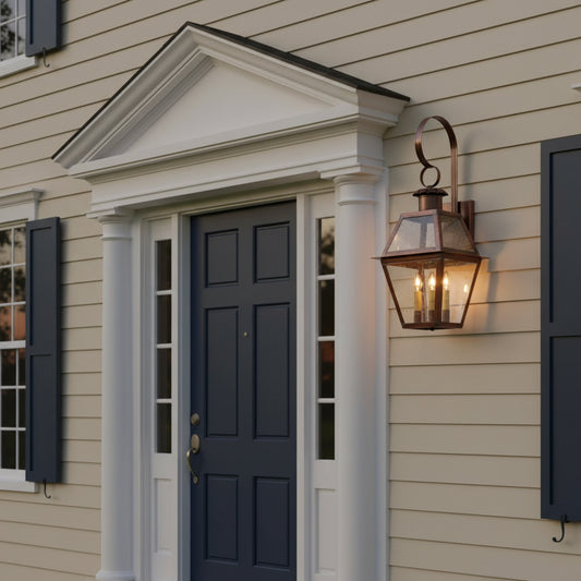An outdoor wall-mounted lantern with a solid antique copper finish and clear glass panels, displaying three lit candles inside on colonial home.