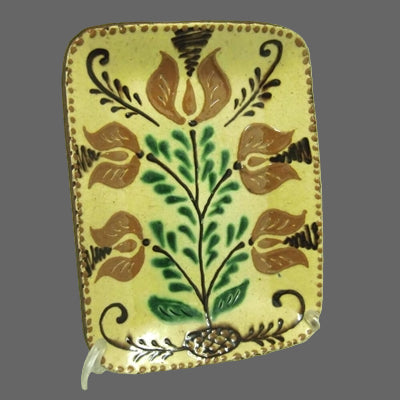 A rectangular ceramic plate with a floral pattern, predominantly yellow with green and brown accents, displayed on a clear stand.