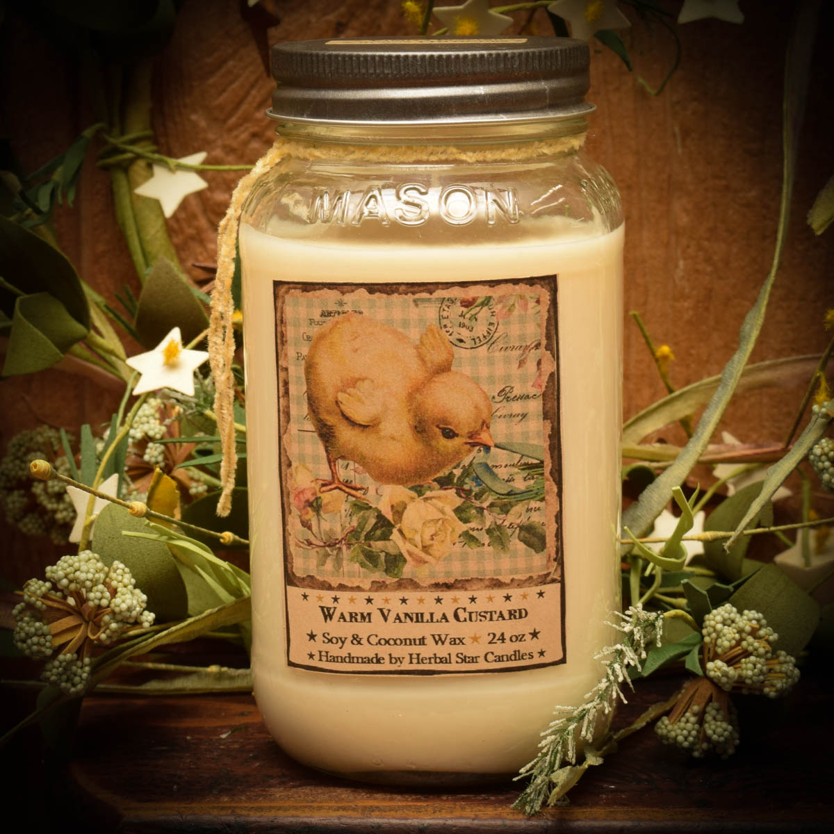 A 24 oz Mason jar candle with a 'Vanilla Custard' label and a decorative vintage-style lid, surrounded by greenery and flowers.