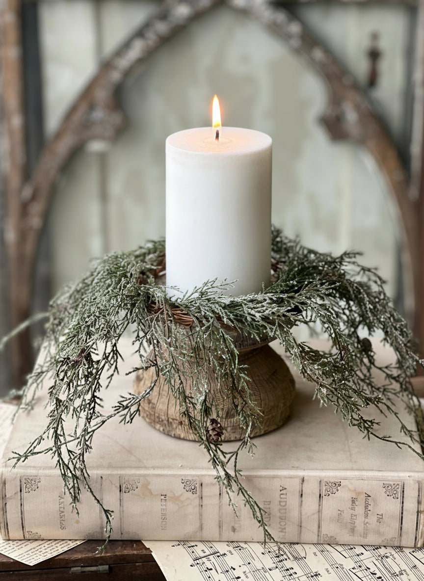 A candle ring featuring greenery and pine cones, dusted with snow and glitter, displayed around a white candle.
