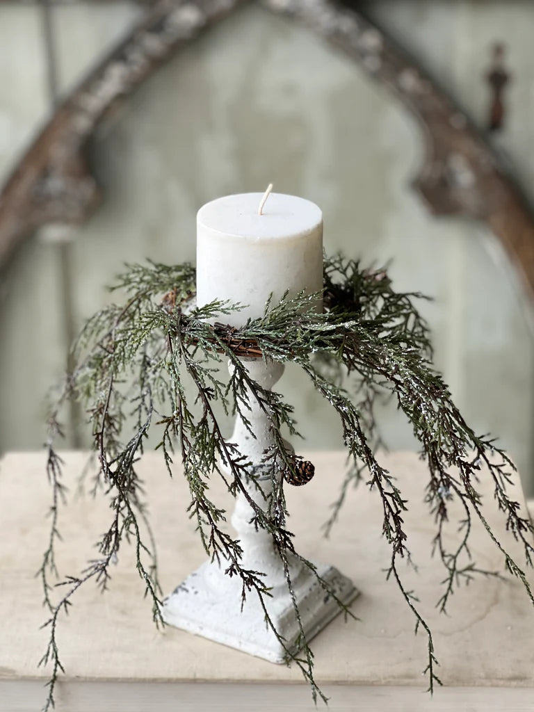 Weeping cedar candle ring with snow and pinecones on a white pedestal holding a white pillar candle.