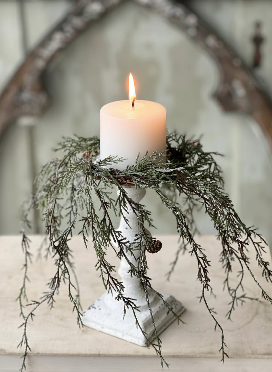 Weeping cedar candle ring with snow and pinecones on a white pedestal holding a white pillar candle.