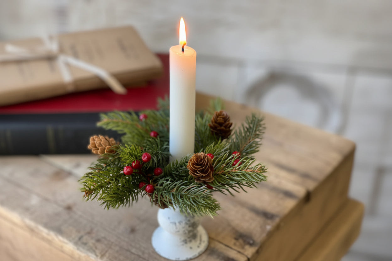 A candle ring with white spruce branches, red berries, and pine cones, displayed around a white candle, placed on a wooden surface.