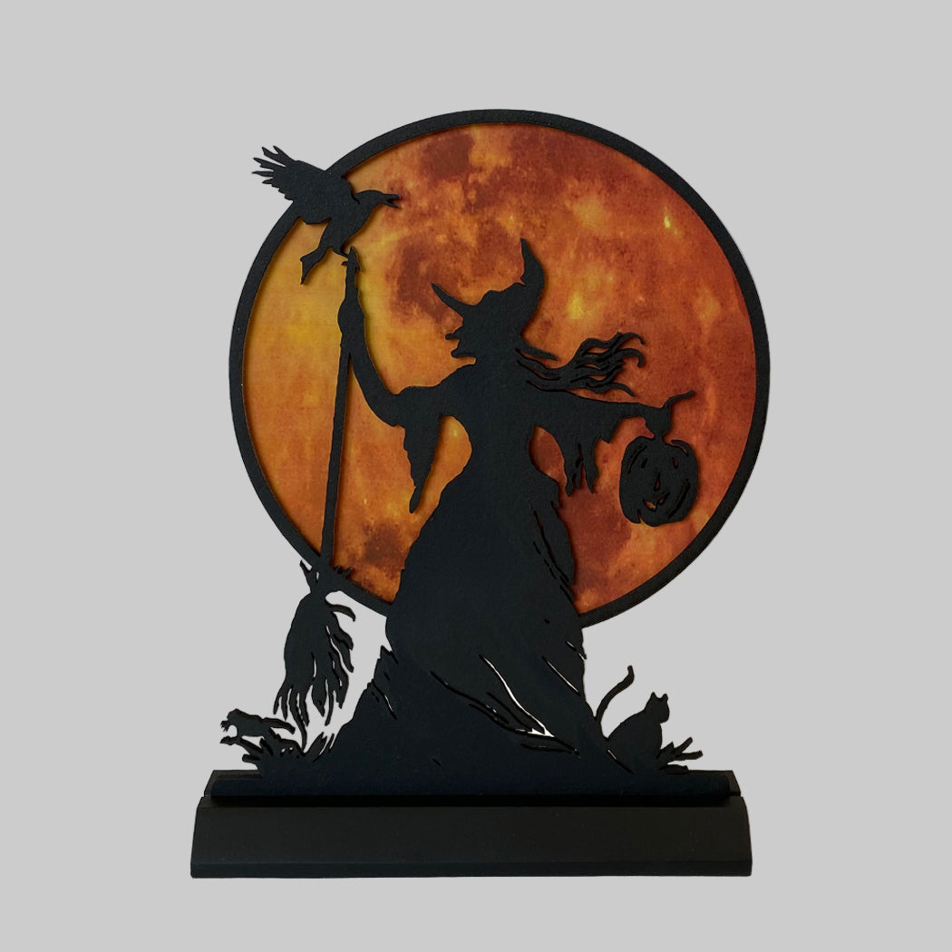 A silhouette of a witch with a broom, cat, and pumpkin, set against an orange and black moon background, designed as a Halloween tabletop ornament.