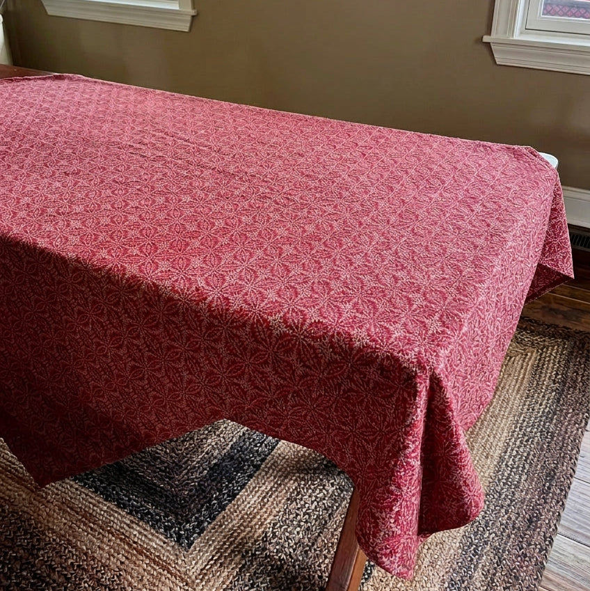 A red patterned tablecloth draped over a wooden dining table in a home setting, styled with a decorative tray, a black lantern, and a small figurine. The background features a beige wall, windows, and a potted green tree.