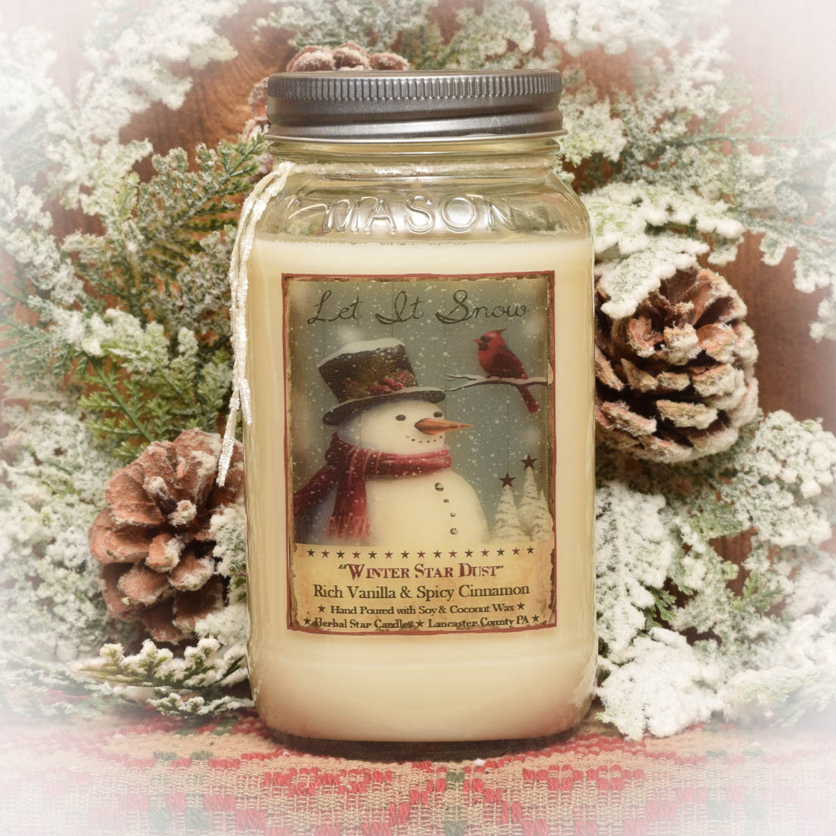 A mason jar candle with a label featuring a vintage-style snowman, pine cones, and the scent marked as 'Winter Star Dust'. The candle is placed in front of a backdrop of pine branches and decorative fabric.