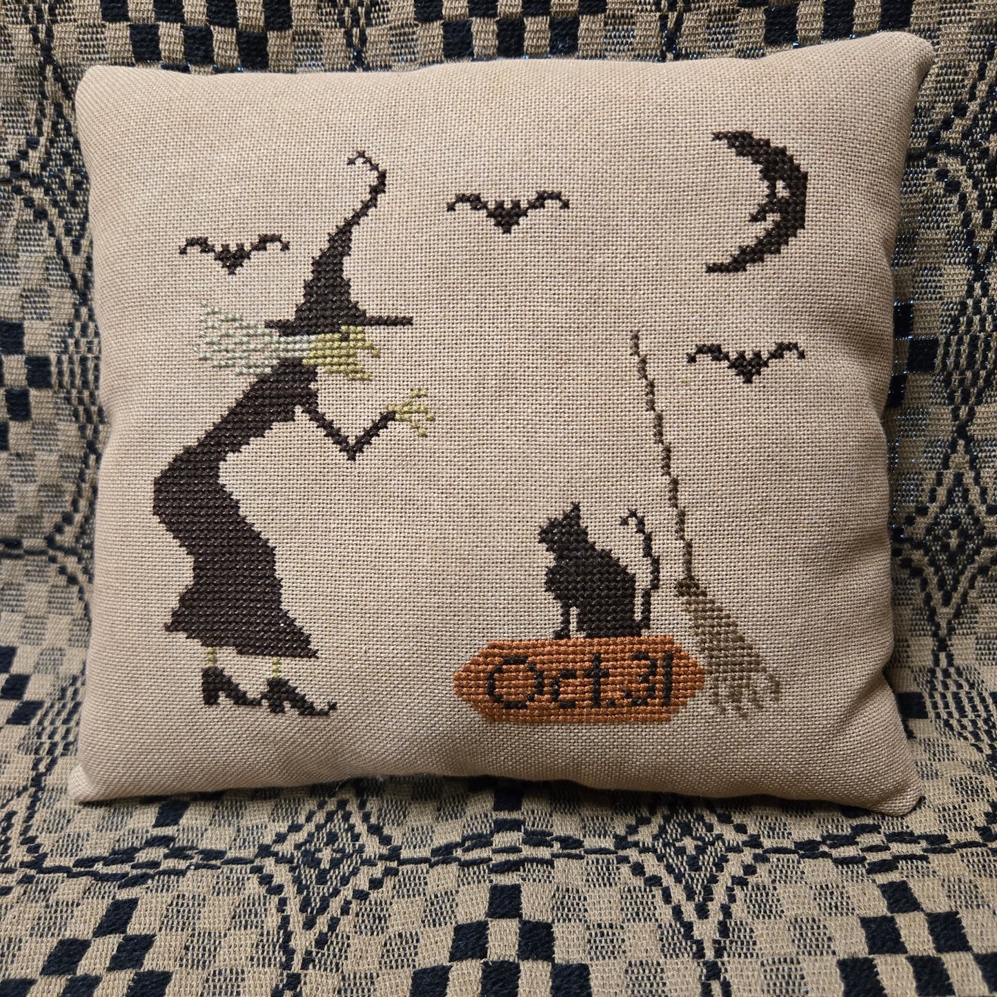 A square beige needlepoint pillow featuring a stitched design of a witch, a cat on a broom, bats, a crescent moon, and the date 'Oct 31' in orange, resting on a blue and white checkered couch.