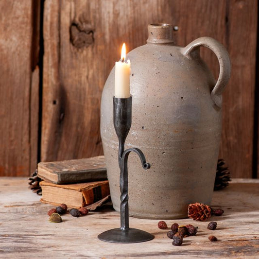 A wrought iron candle holder in an antique finish, with a candle lit, placed on a wooden surface next to a ceramic jug and pine cones.