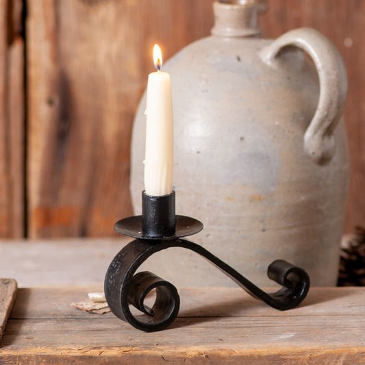 A wrought iron curled candle holder in black color, placed on a wooden surface with a candle lit inside it, set against a rustic backdrop with a jug and wooden elements.