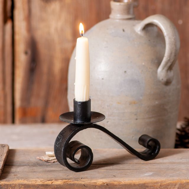 A wrought iron curled candle holder in black color, placed on a wooden surface with a candle lit inside it, set against a rustic backdrop with a jug and wooden elements.