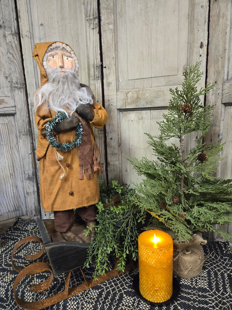 A handcrafted clay figurine of Santa wearing brown pants, a brown coat, and a festive wreath, standing next to a decorative evergreen tree and a lit candle, placed on a patterned textile.