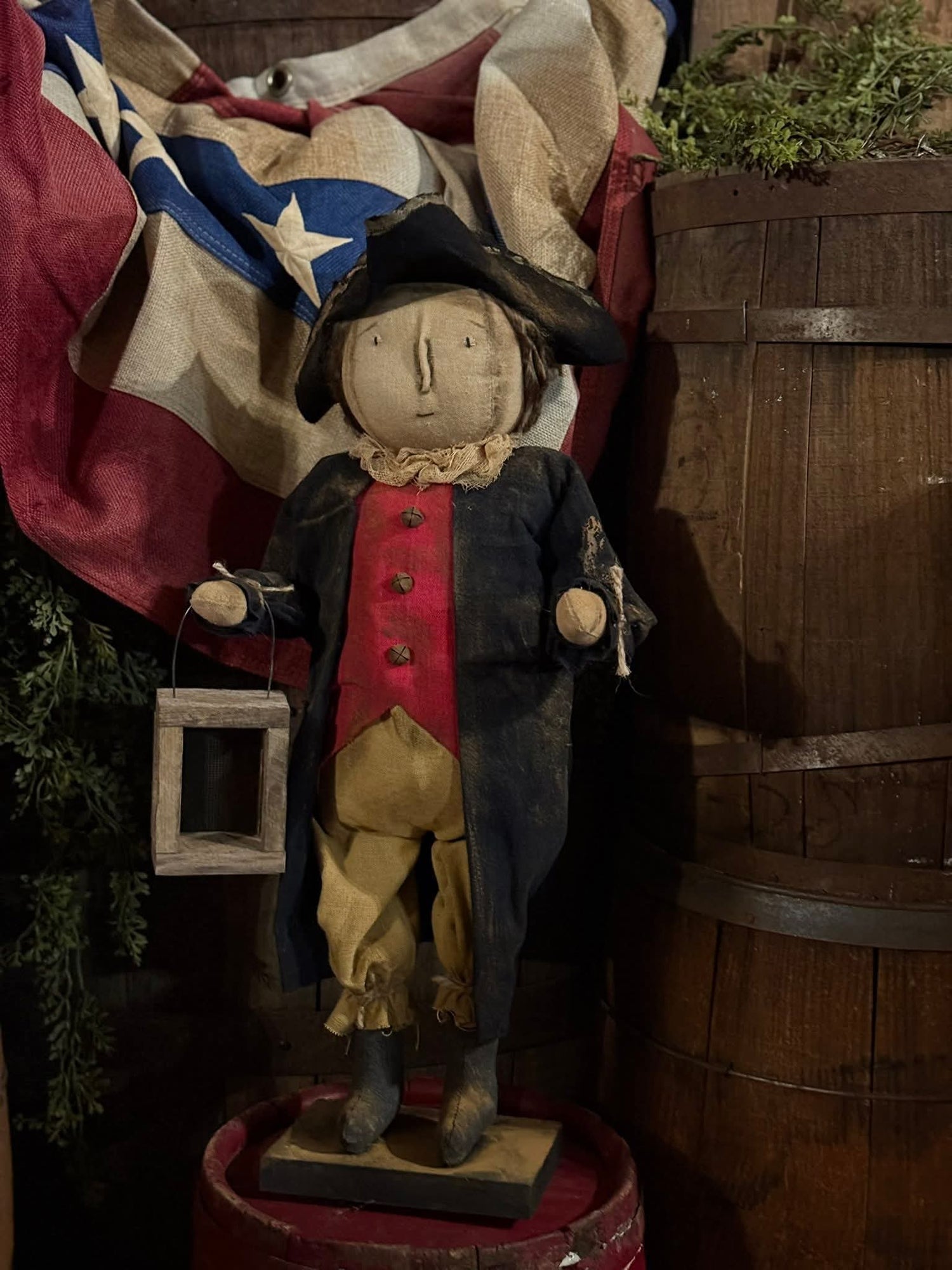 Paul Revere doll holding a wood lantern in front of a barrel and flag.