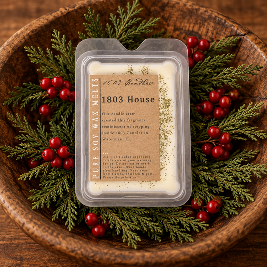 A packaged soy wax melt labeled '1803 House' by 1803 Candle Co., placed in a wooden bowl with evergreen branches and red berries.