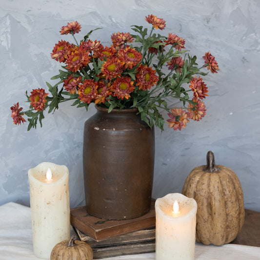A burnt orange strawflower pick placed in a ceramic vase with autumn-colored flowers, surrounded by candles and a book on a rustic table.