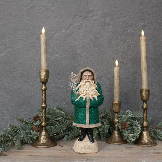 An 11-inch green belsnickel figure with a white beard and a feather branch, standing between two lit candles with a decorative branch in the background.