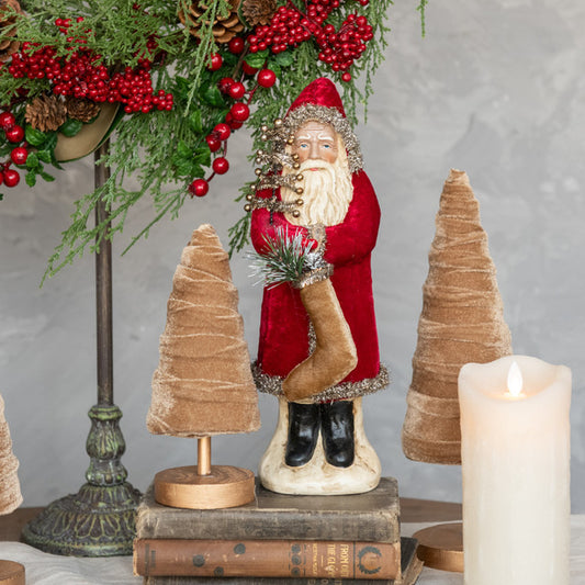 A 12-inch tall wine velvet belsnnickle figure, depicting a Santa-like character, with a glass of wine, seated on a book, with a decorative tree and pinecones in the background, and a lit candle beside it.