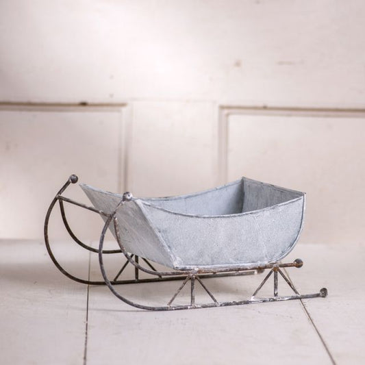 A 13-inch tin sleigh with a rustic white wash finish and solid steel runners, displayed on a hearth or tabletop.