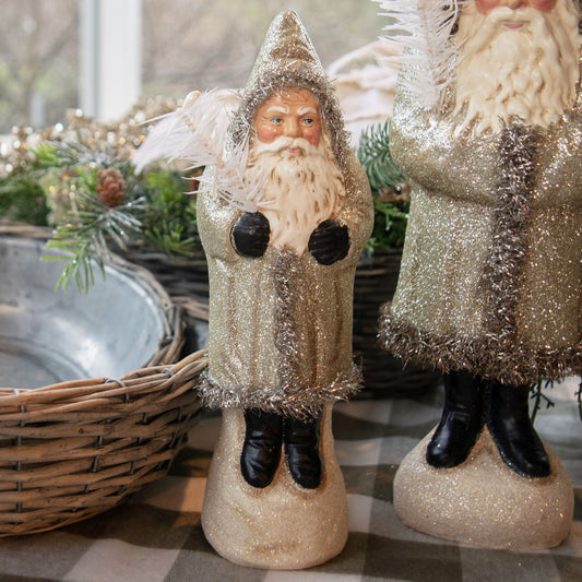 Two champagne glitter Belsnickle figurines with feather twigs, one standing upright and the other sitting down, set against a holiday-themed backdrop with Christmas decorations.