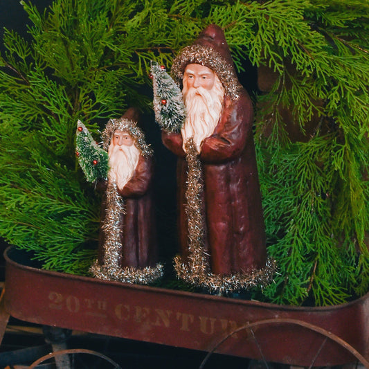 Two red belsnickels holding bottlebrush trees stand in old wagon with green wreath behind them.