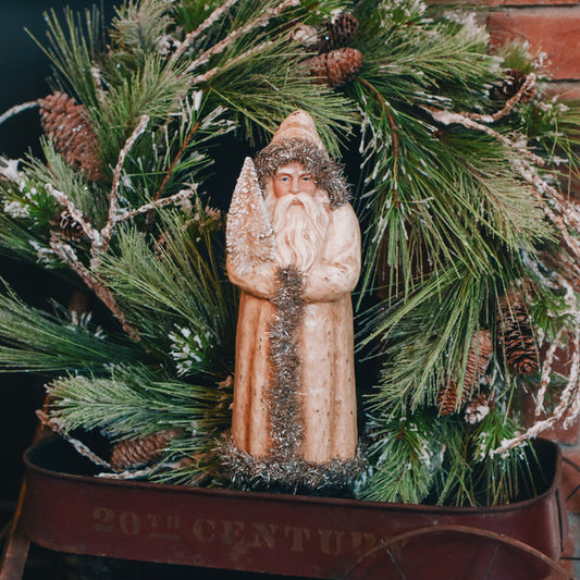 A beige belsnickle figurine with a white beard and hat, standing amidst green pine branches and brown holly leaves.