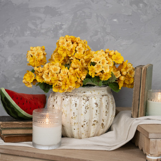 A artificial yellow geranium bush in a white vase with real yellow flowers, placed on a wooden table with a slice of watermelon and two lit candles.