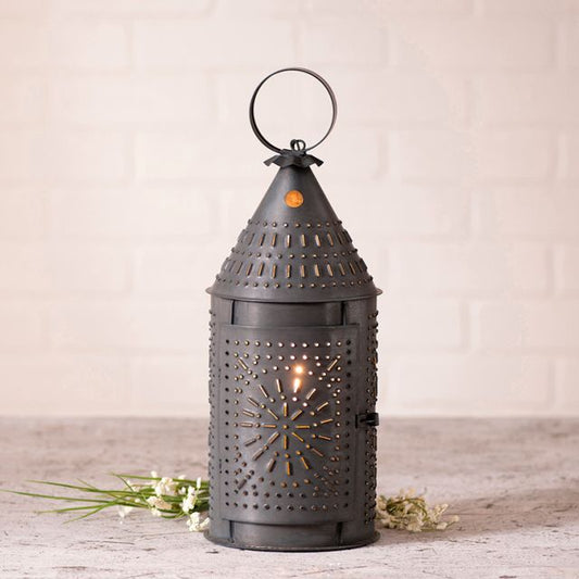 A black metal Revere lantern with a punched pattern design, illuminated from the inside, placed on a grey surface with a white brick wall background.