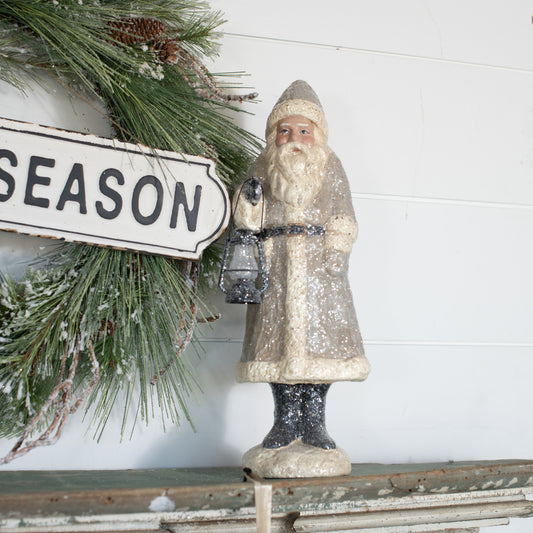 A taupe-colored belsnickel figurine holding a lantern, standing in front of a 'SEASON' sign, with holiday decorations in the background.
