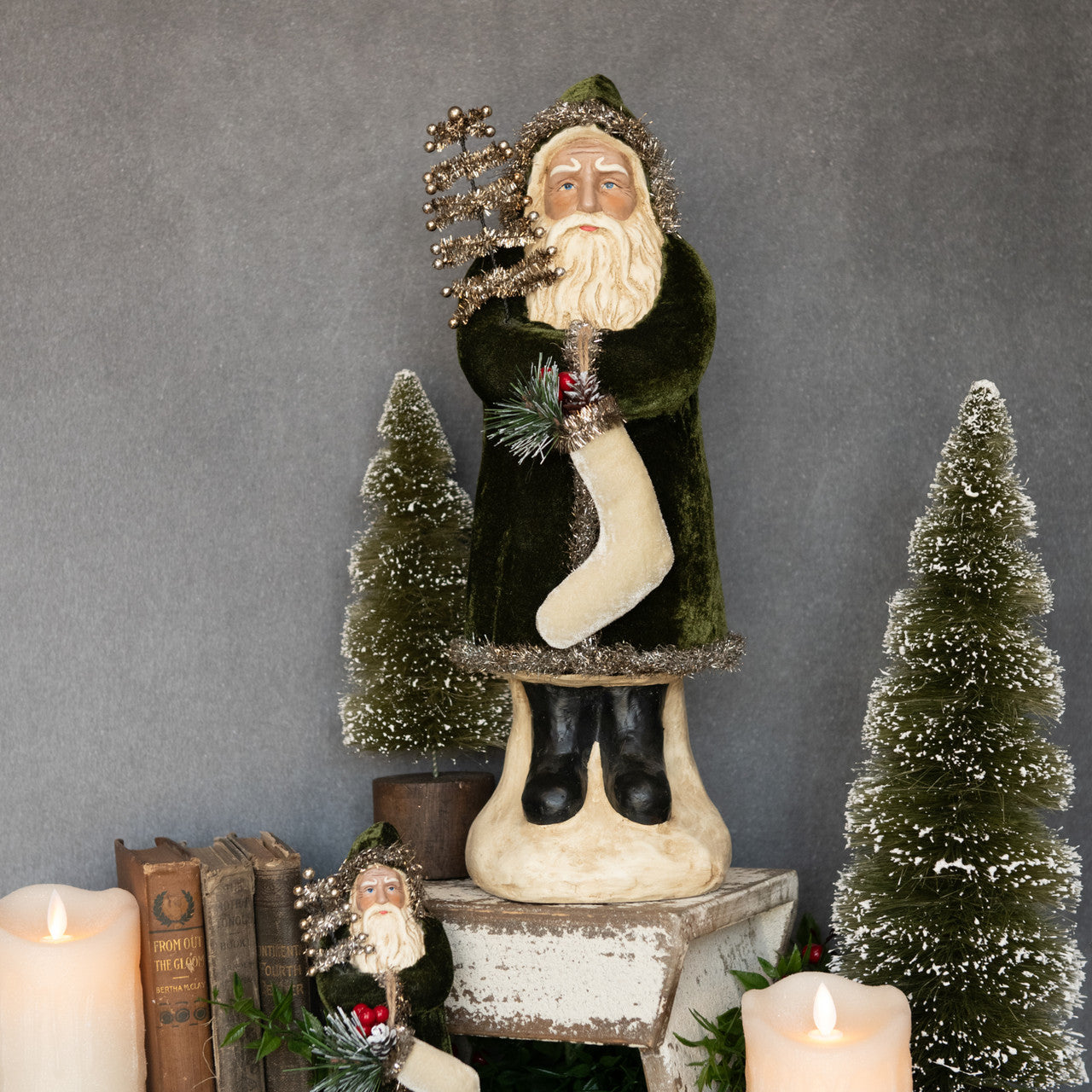Decorative figure of a man holding a tree and a stocking, surrounded by candles and small trees on a gray background.