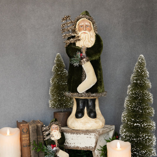 Decorative figure of a man holding a tree and a stocking, surrounded by candles and small trees on a gray background.