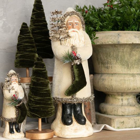 A pair of belsnickle figurines made of oatmeal velvet, one large and one small, with the large one holding a Santa hat, standing beside a decorative potted plant and a wooden object, all part of a holiday decoration setup.