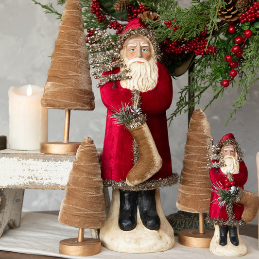A 16.5 inch tall wine velvet belsnickel figurine, depicting a Santa-like character, with a red jacket, white beard, and a brown belt, standing next to a decorative tree.